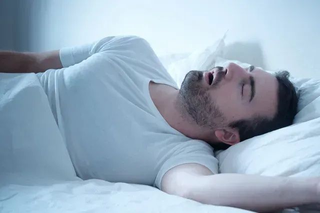 Man sleeping on a white bed, mouth open, snoring.