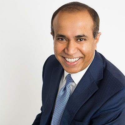 Man with dark hair smiling in a blue suit and tie, white background.