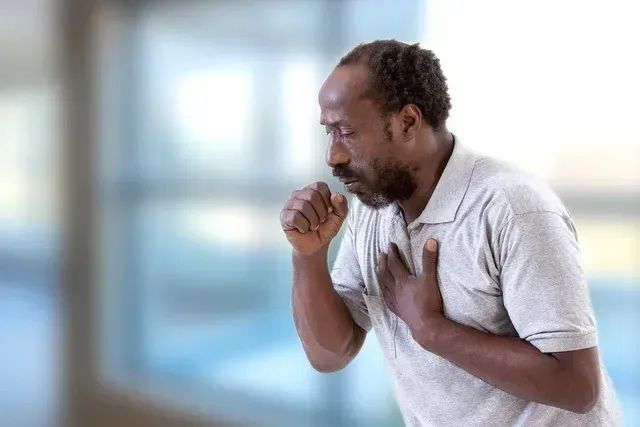 Man coughing, holding chest, wearing gray shirt, blurred background.