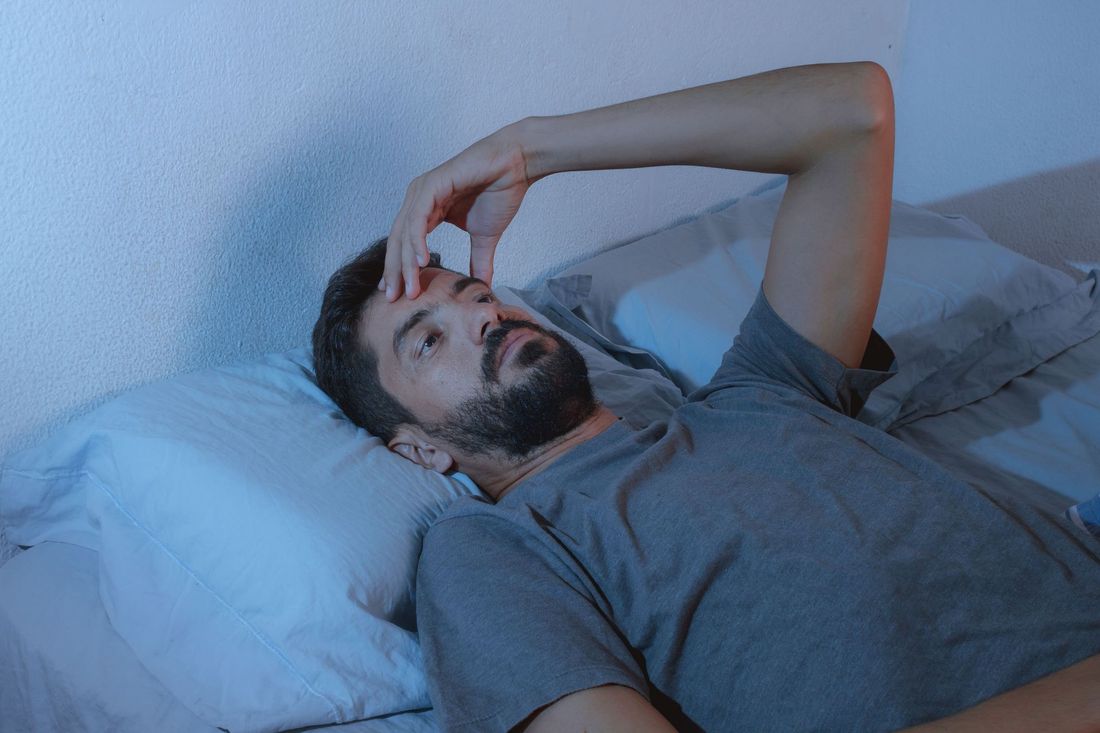 Man in bed, looking stressed, hand on forehead, illuminated by blue light.