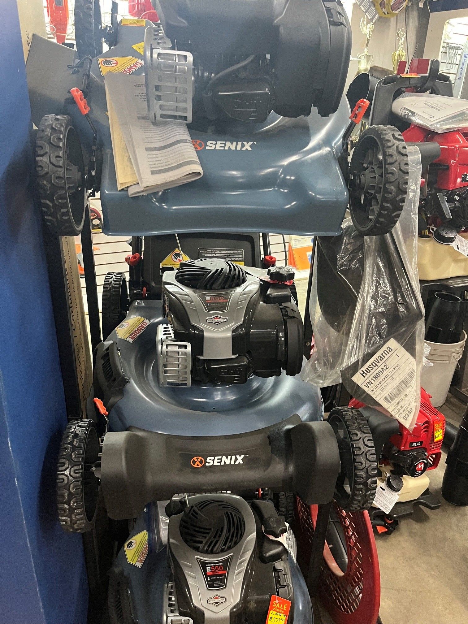 Stacked blue lawn mowers on a store display, with one red mower at the bottom.