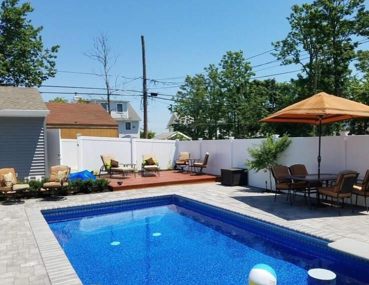 Backyard with pool, deck, and patio furniture. Blue pool, white fence, sunny day