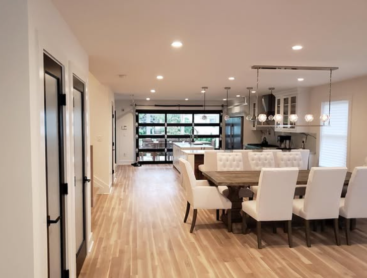 Open-concept dining and kitchen area with wooden floors, a long table, white chairs, and a glass garage door