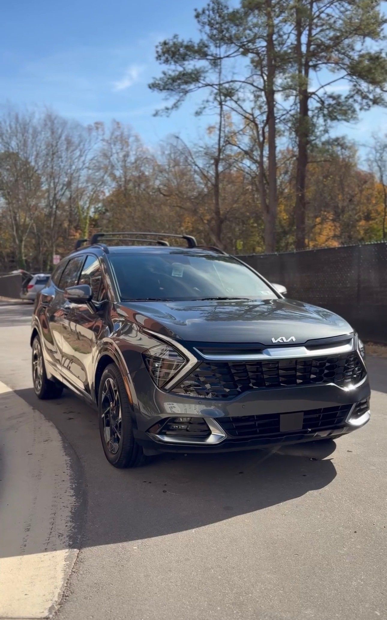 Dark gray Kia Sportage SUV parked on an asphalt road; trees and a blue sky in the background.