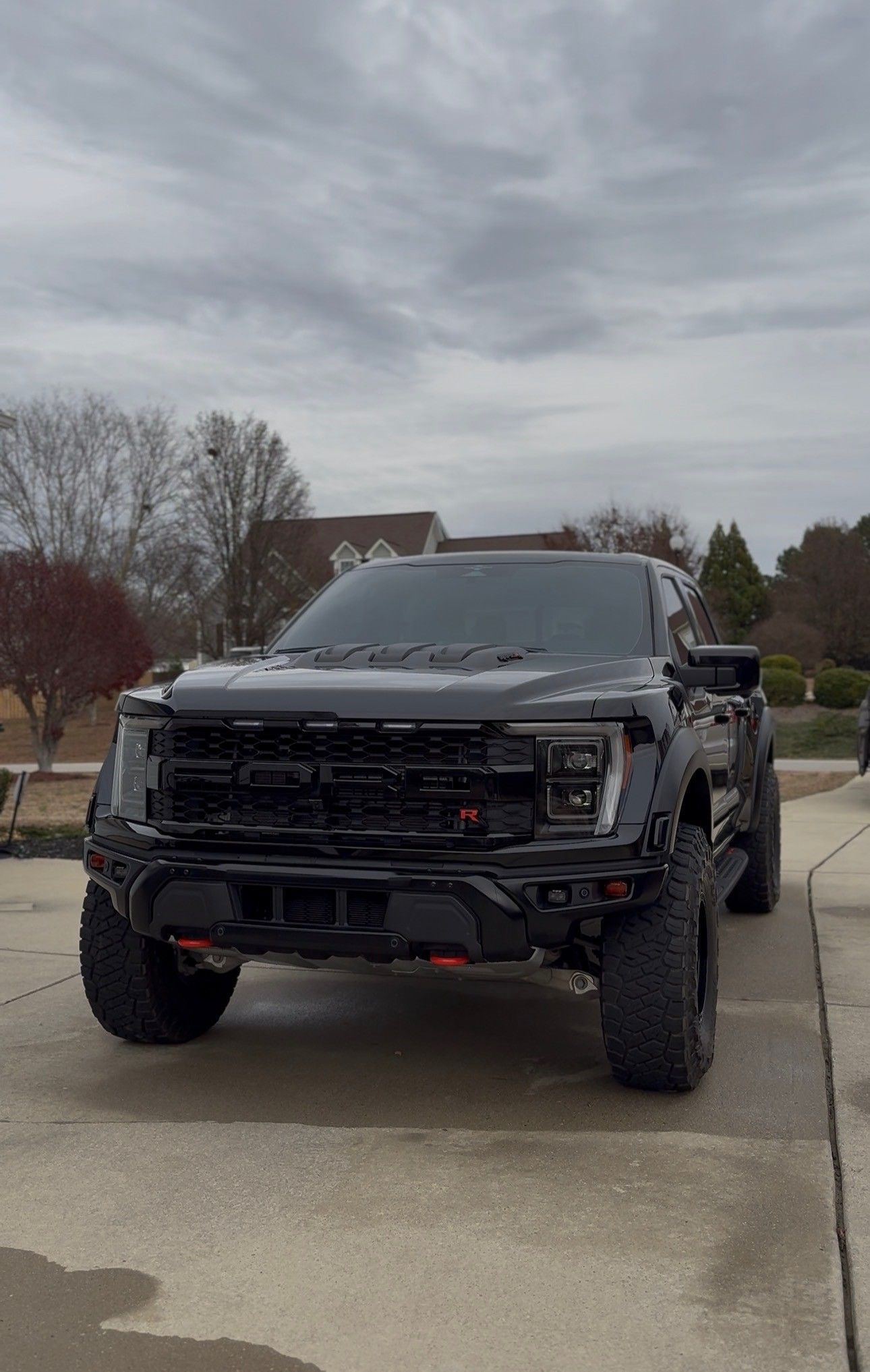 Black Ford Raptor truck parked on a concrete driveway under a cloudy sky.
