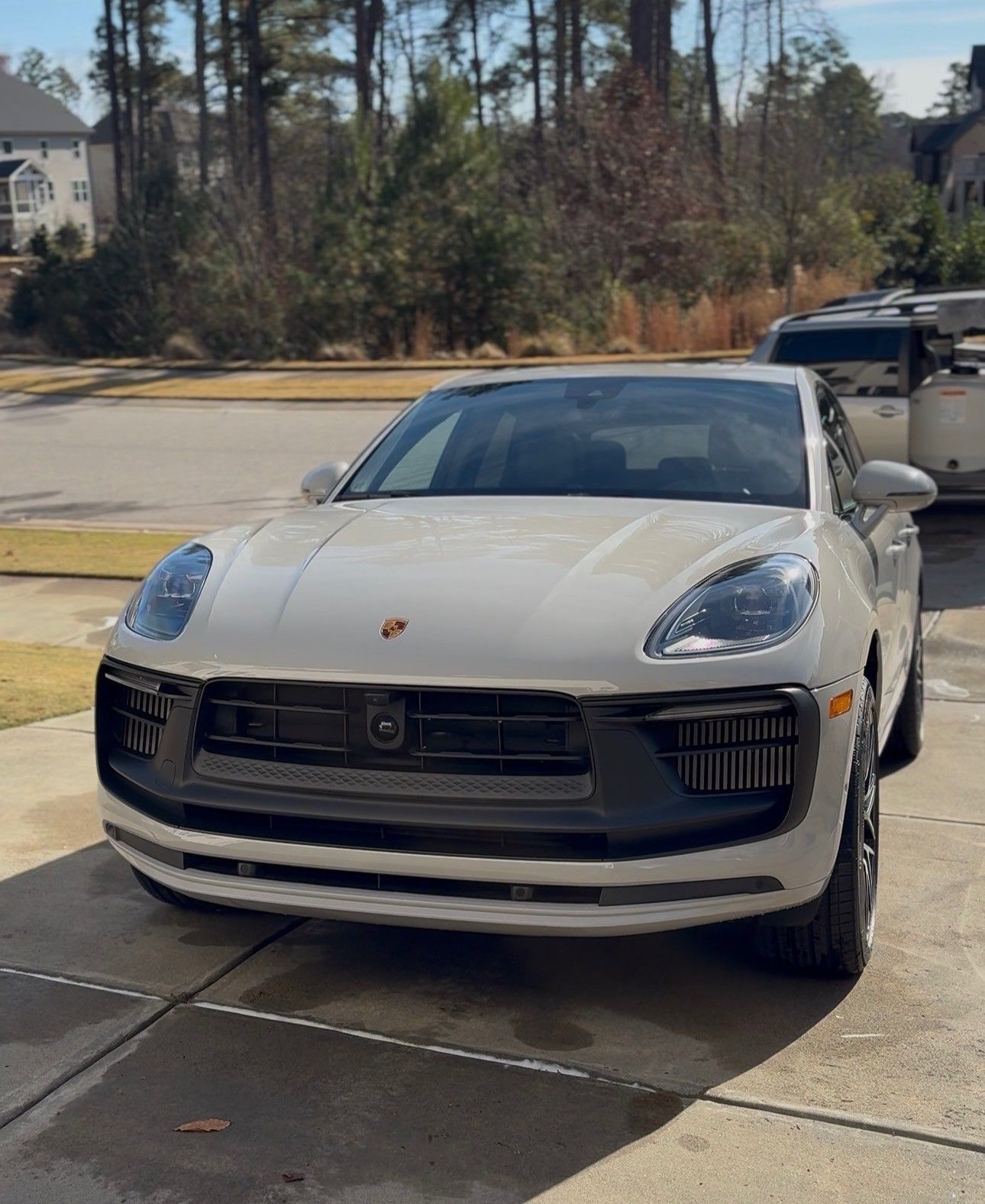 White Porsche SUV parked on a paved driveway in front of a house on a sunny day.