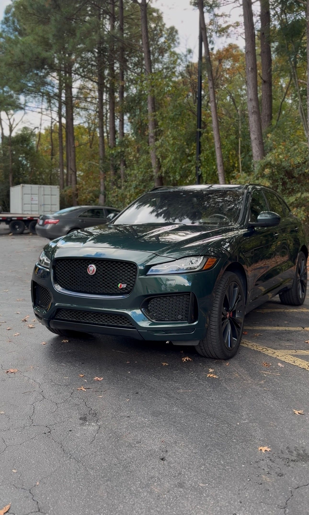 Green Jaguar SUV parked outdoors. Black grill, wheels. Trees in background.
