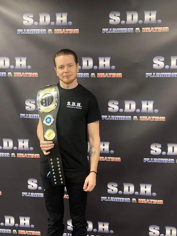 Man in black shirt and pants holds championship belt in front of a banner for plumbing and heating.