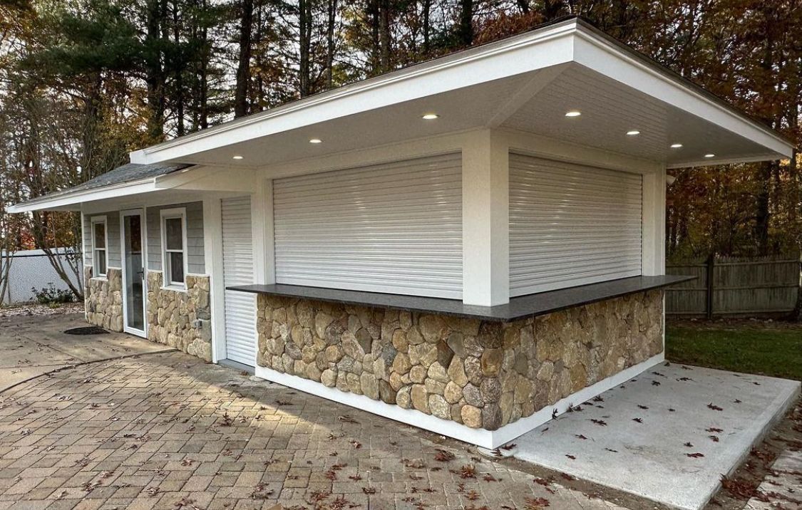 Modern small building with stone base, white roof overhang, and large shuttered windows in a wooded area