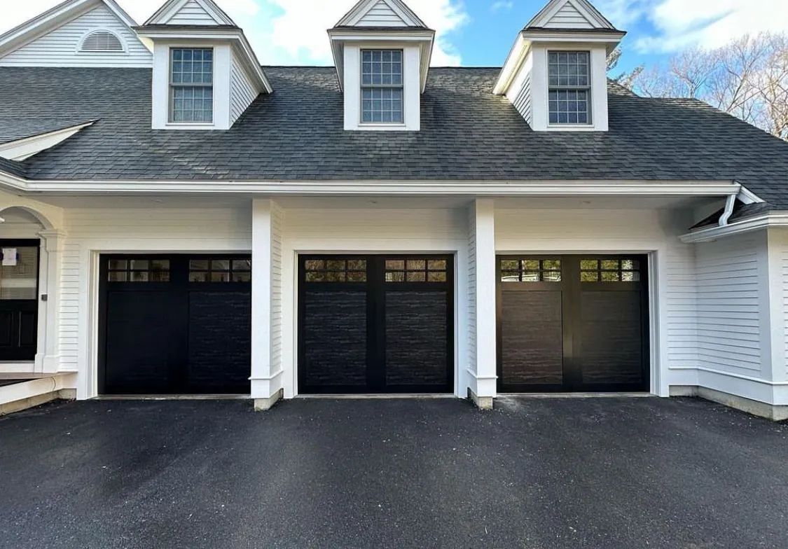 Three-car garage with black doors and white siding beneath dormer windows on a paved driveway