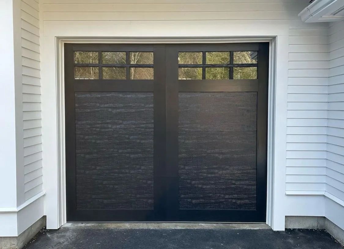 Black double garage doors with small windows, set in a white house exterior.