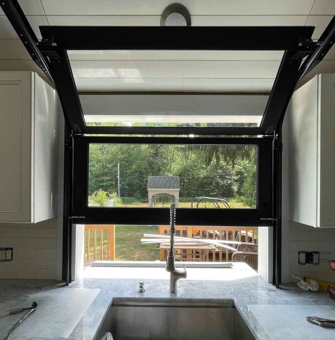 Modern kitchen sink under a large window, looking out onto a bright backyard deck and trees