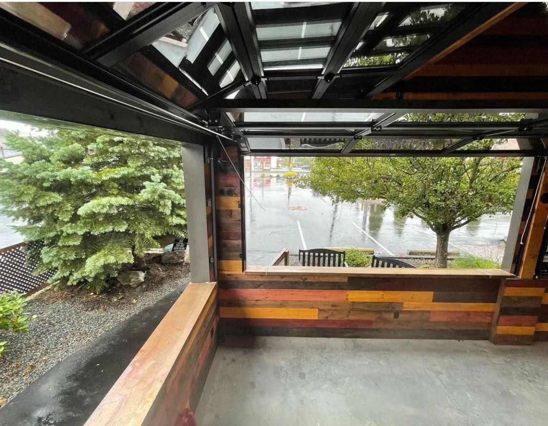 Covered patio overlooking a rainy waterfront, with wooden benches and a green tree nearby.