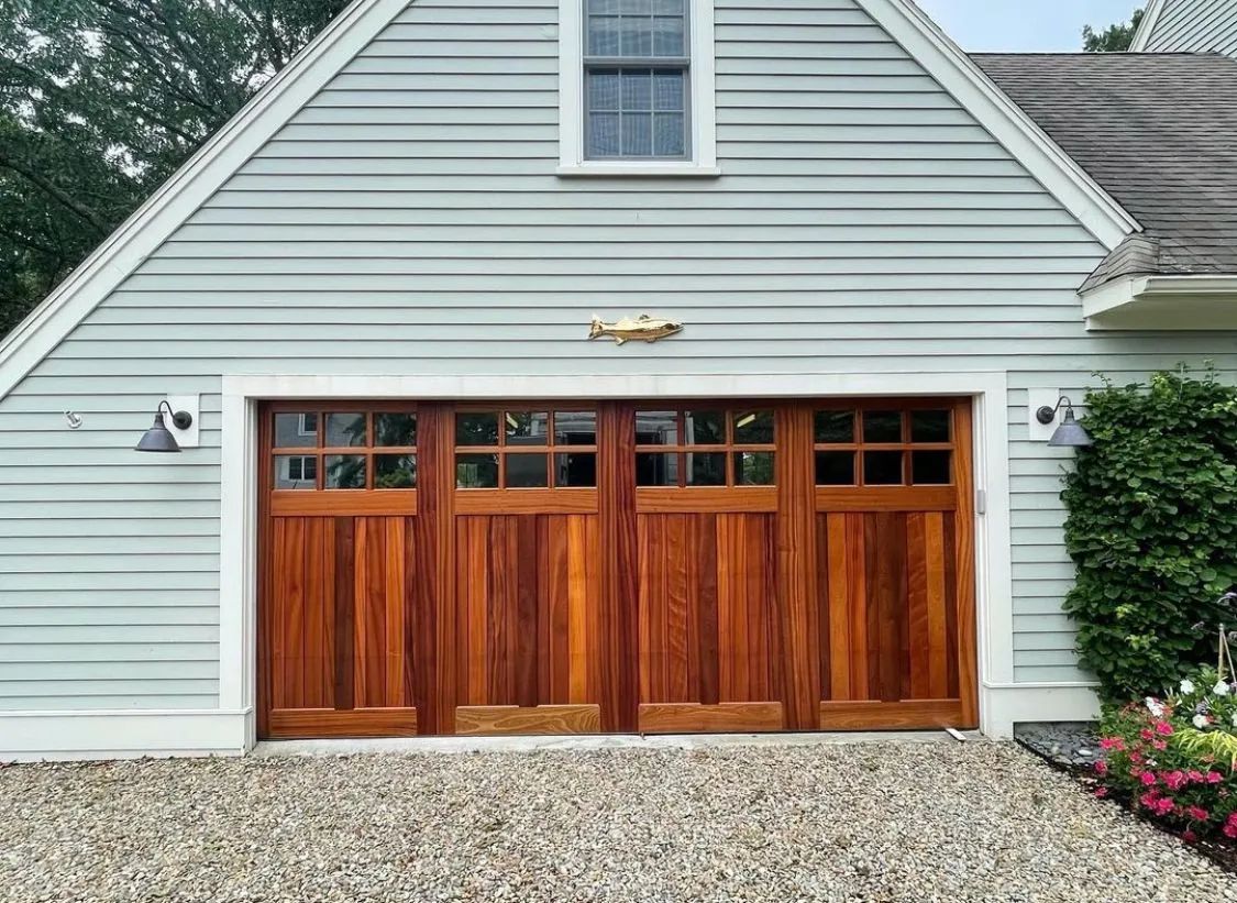 Gray garage with four wooden doors and a small upper window, set against a gravel driveway and shrubs