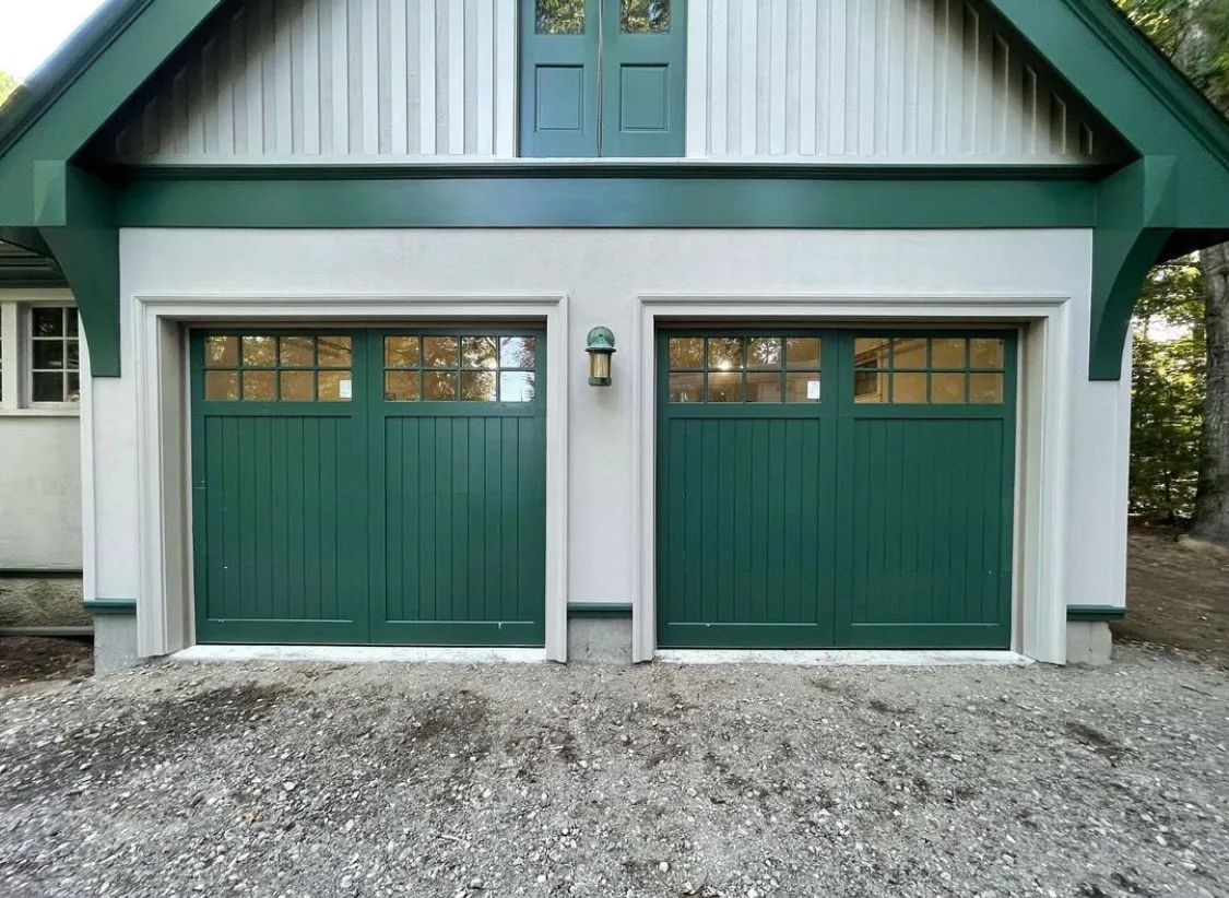 Front view of a white garage with two green doors and a green gable roof over gravel driveway
