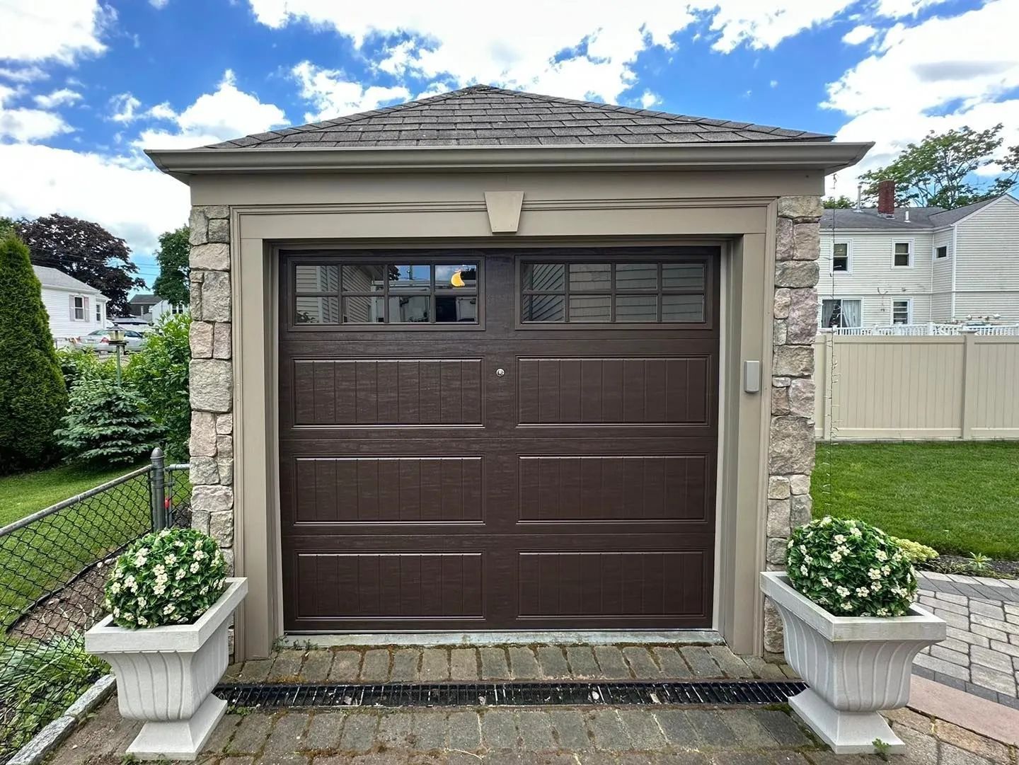 Brown garage door with small windows on a stone house, framed by white planters and a lawn.