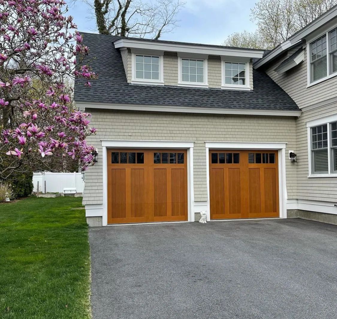 Two-car garage with wood doors on a gray house beside a blooming pink tree and gravel driveway
