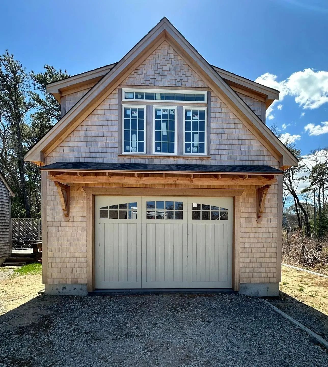 Small beige garage with a gabled roof, white doors, and upstairs windows under a blue sky.