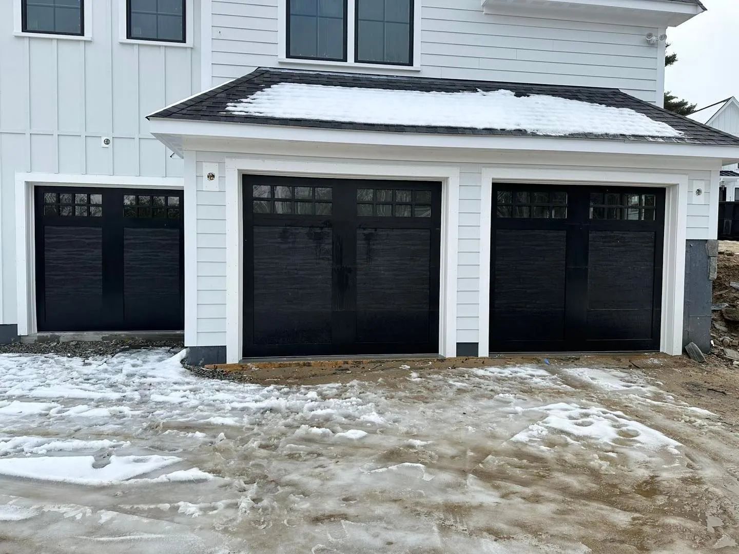 Snow-covered driveway in front of a house with three black garage doors and white siding