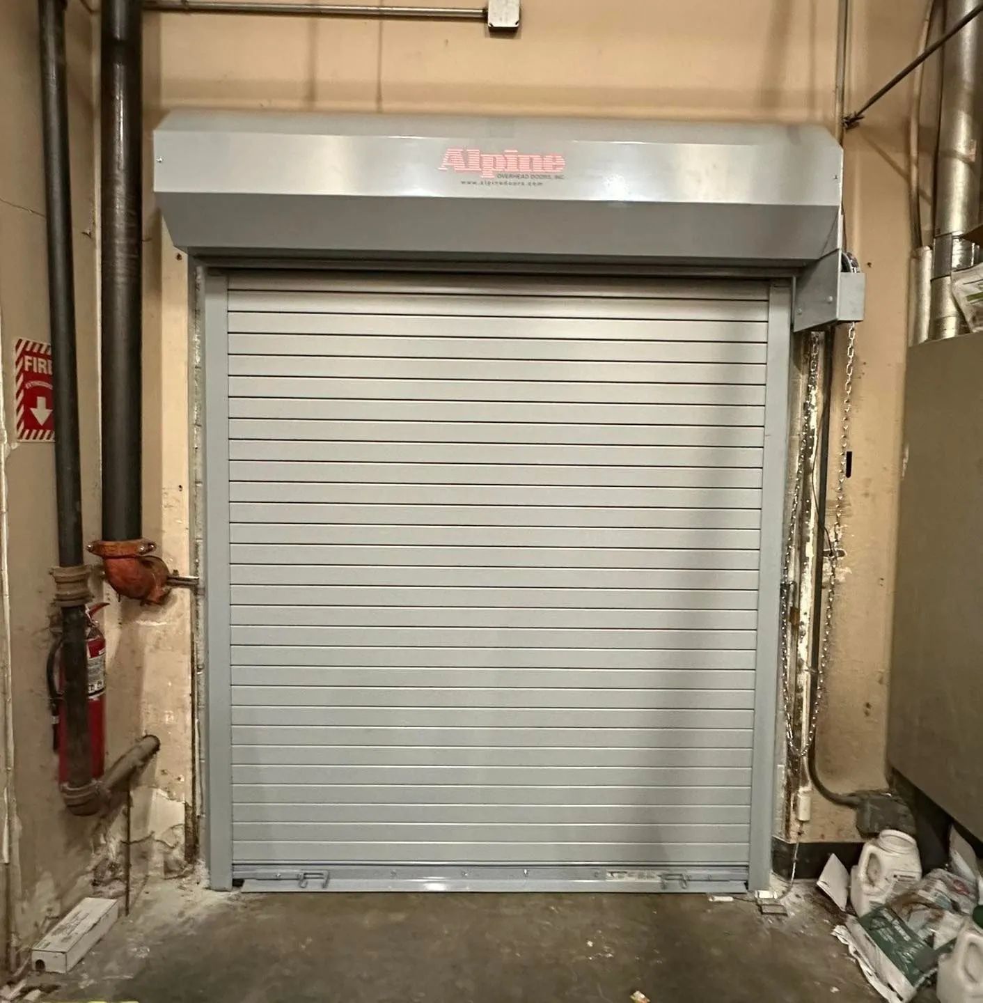 Closed silver roll-up warehouse door in an industrial hallway, with debris along the floor.