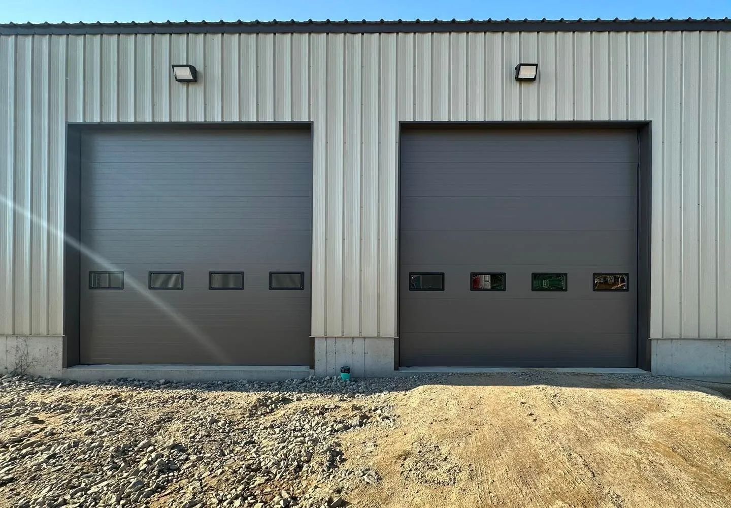 Two gray roll-up garage doors on a metal building, with gravel in front and sunlight glare on the left.