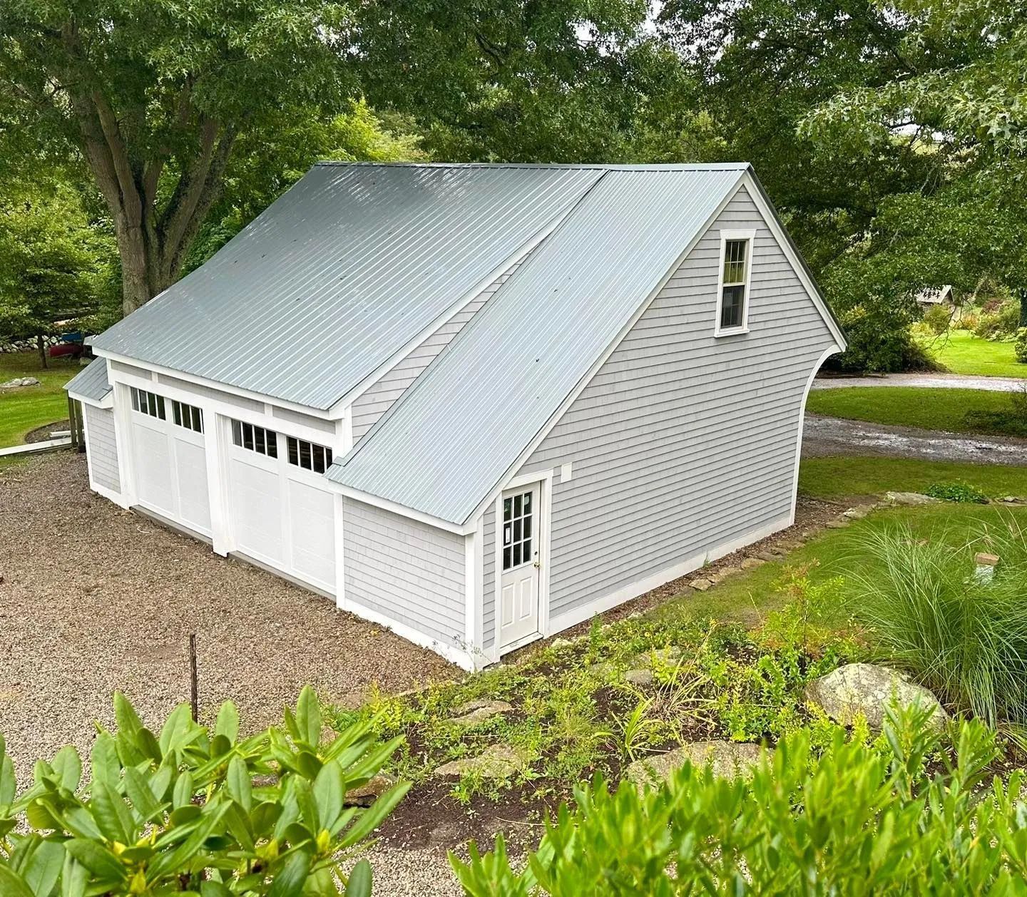 White garage with silver metal roof and dormer window, surrounded by trees and greenery.
