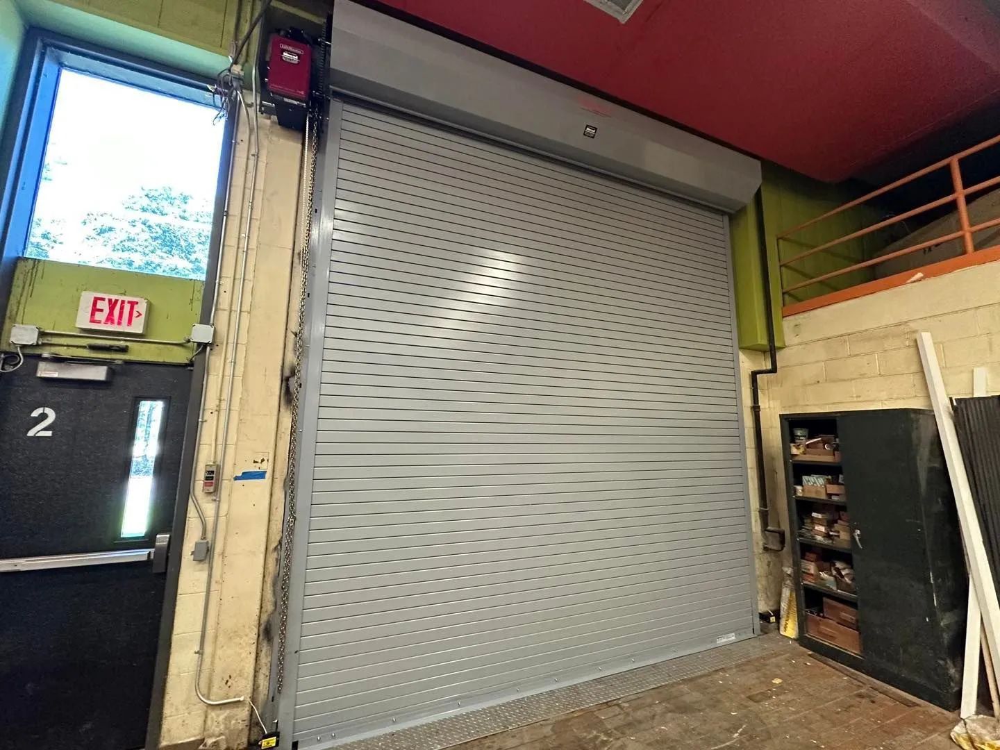 Large gray metal roll-up door in an industrial interior beside an exit door and storage shelves.