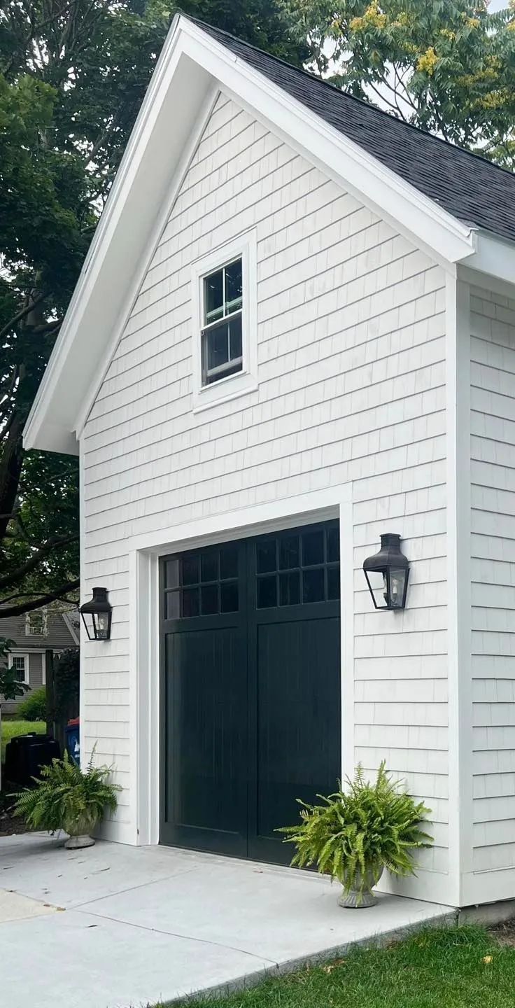 White garage with dark green doors on a house exterior, set beside a driveway and potted plants.