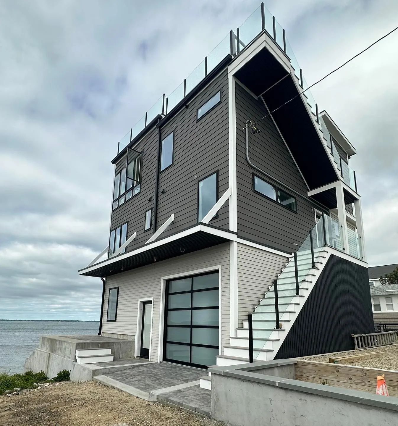 Modern seaside house with dark siding, glass garage, and exterior staircase under cloudy skies
