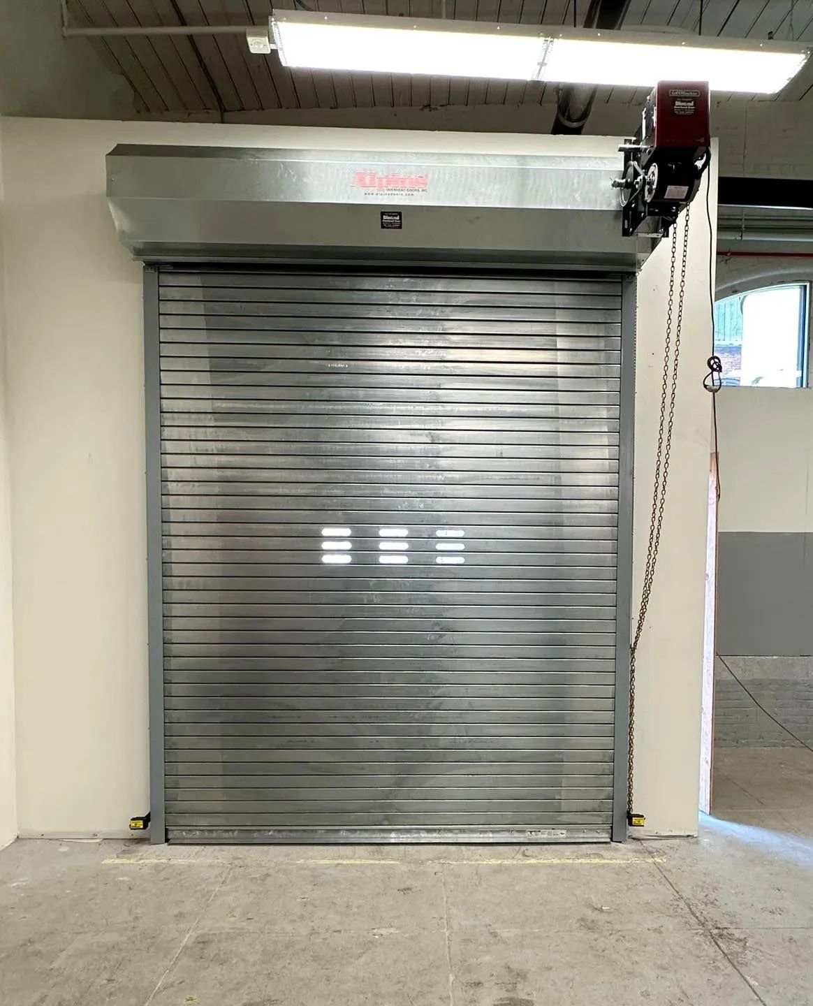 Metal roll-up industrial door with a chain hoist, closed in a warehouse or garage interior.