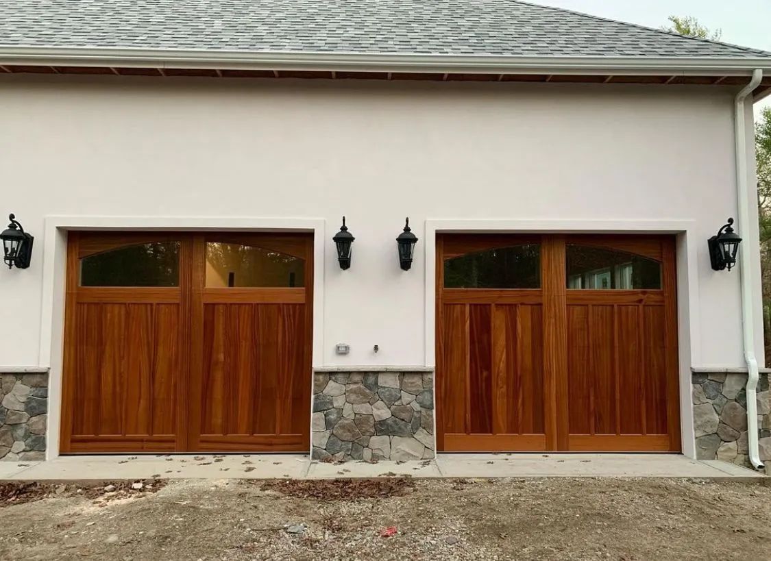 Stone garage with two wooden double doors and black wall lanterns on a white house exterior