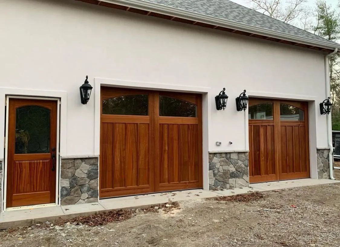 White house exterior with wooden garage doors, side door, stone base, and wall lanterns