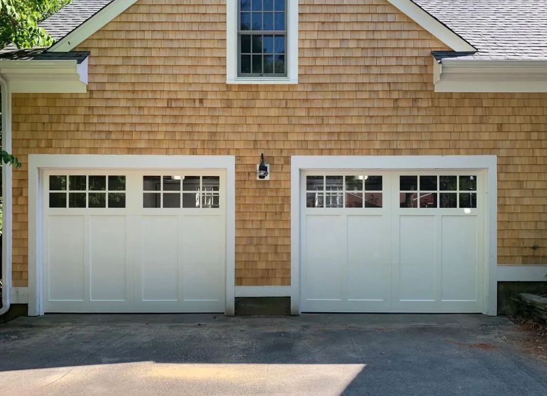 Two white garage doors on a tan shingle-sided house with a driveway in front