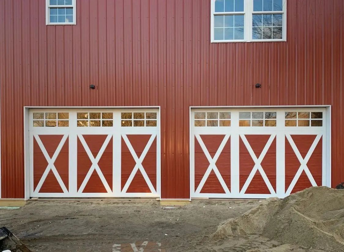 Red barn with two white X-braced garage doors and upper windows, set on a dirt foundation