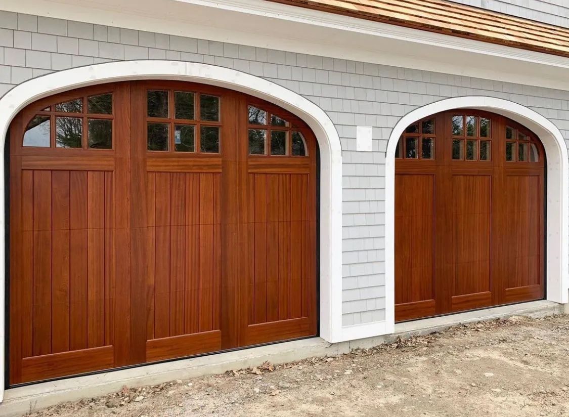 Two wooden garage doors with arched white trim on a gray-sided house exterior
