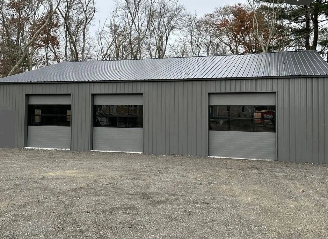 Gray metal garage building with three roll-up doors on a gravel lot, surrounded by trees.