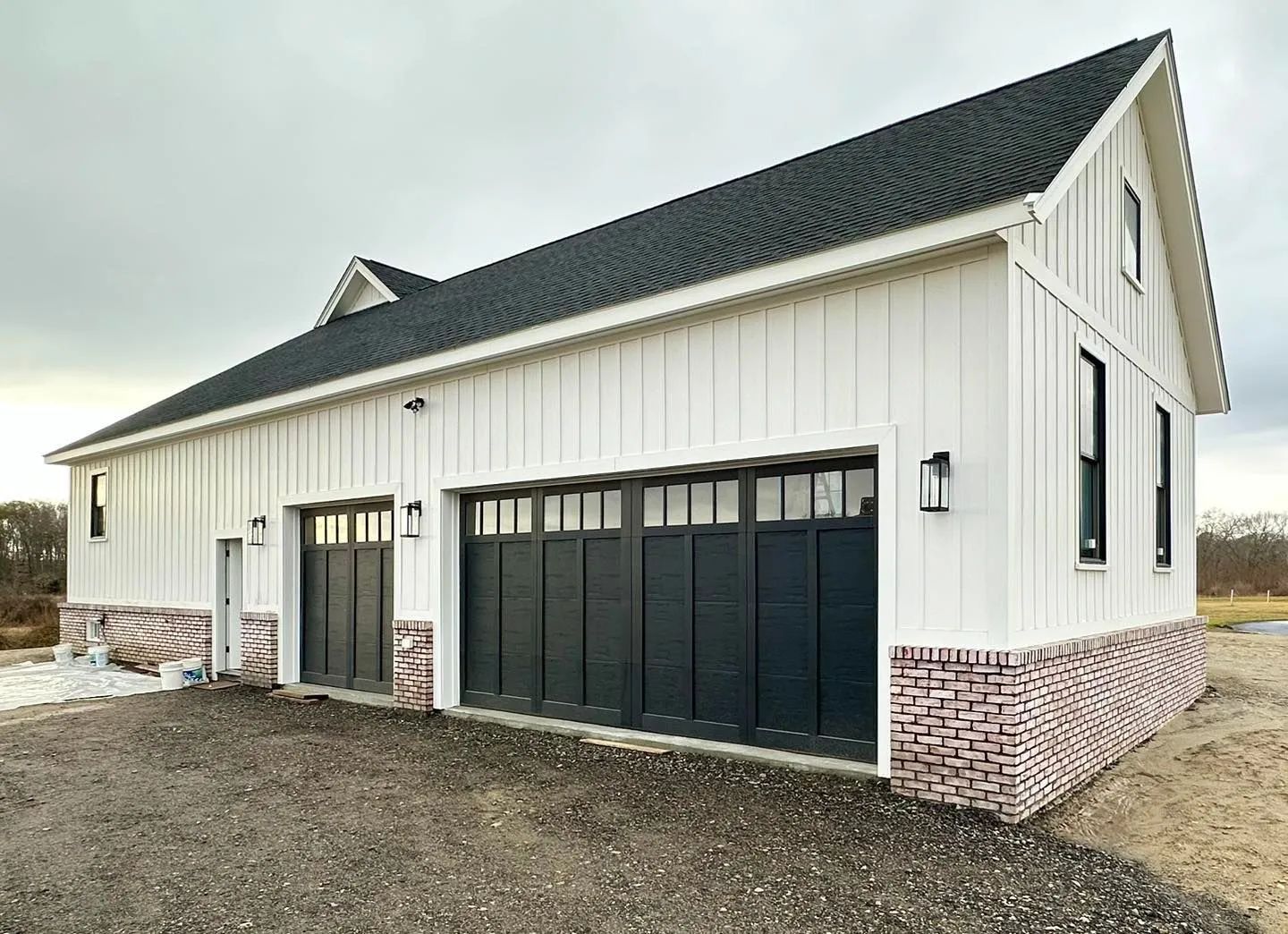 White barn-style garage with black doors, brick base, and shingled roof on a gravel lot.