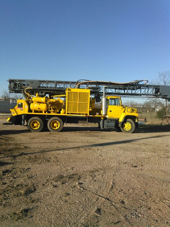 Yellow drilling rig truck on a gravel lot under a blue sky.