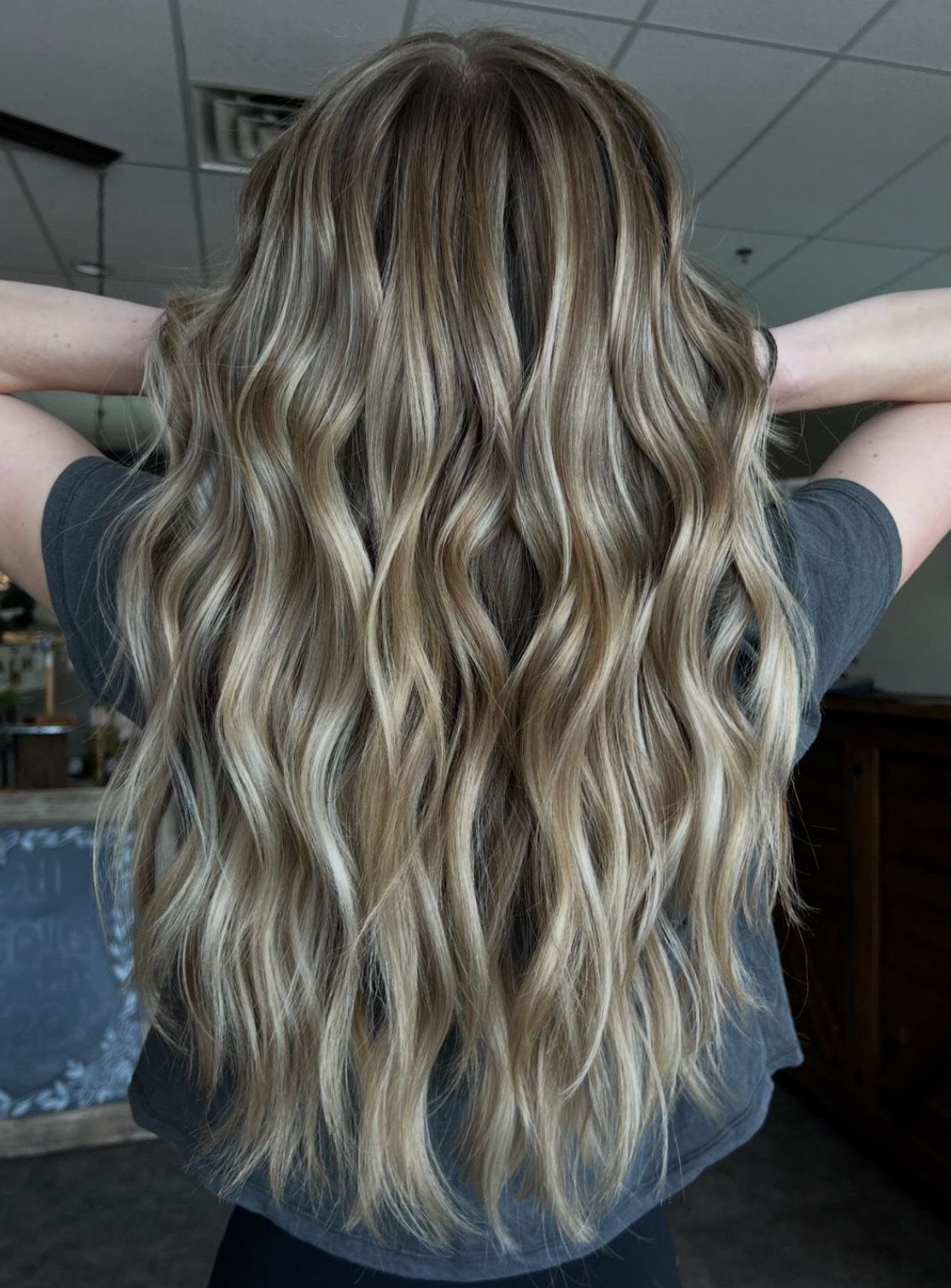 A woman with long blonde hair is standing in a room with her hands behind her head.