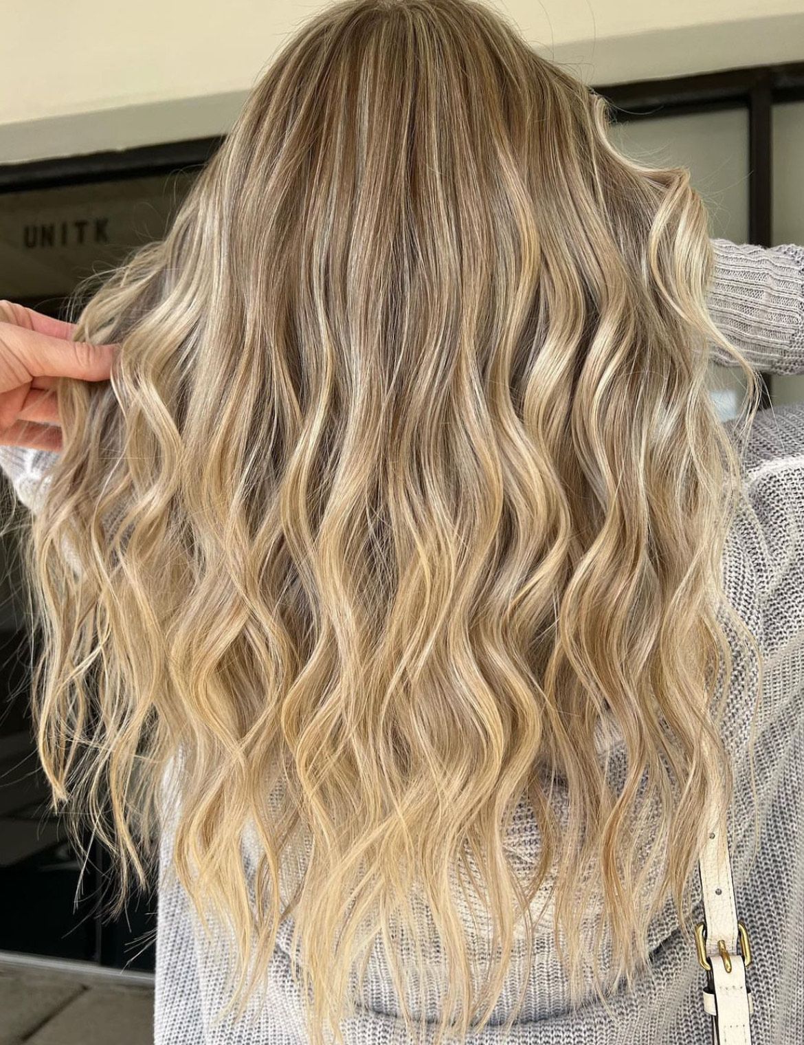 A woman with long blonde wavy hair is wearing a gray sweater.