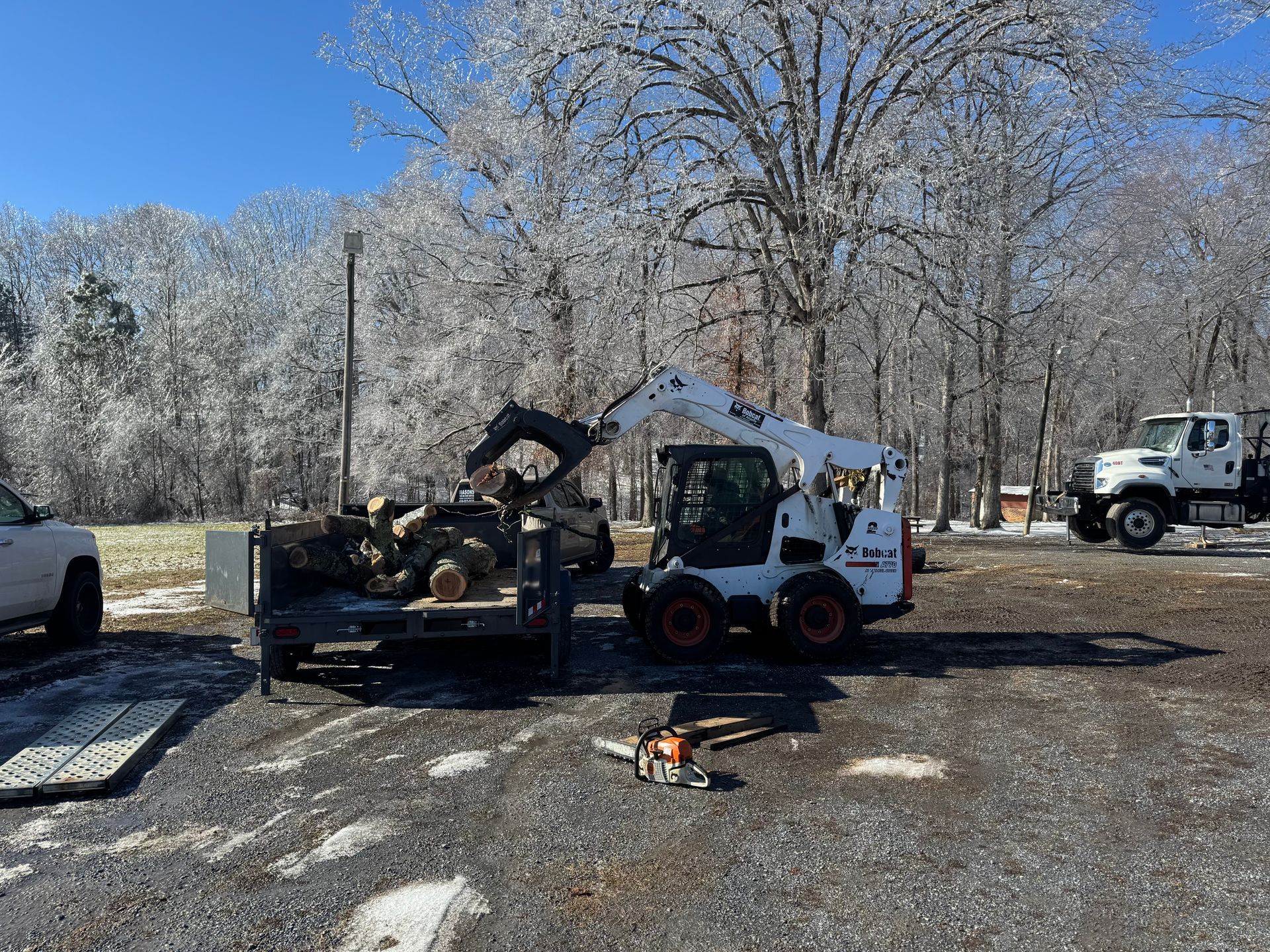 A bobcat is carrying logs on a trailer in a parking lot.