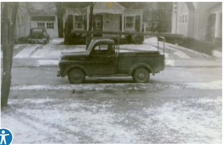 A vintage pickup truck parked on a snow-covered street in front of houses.