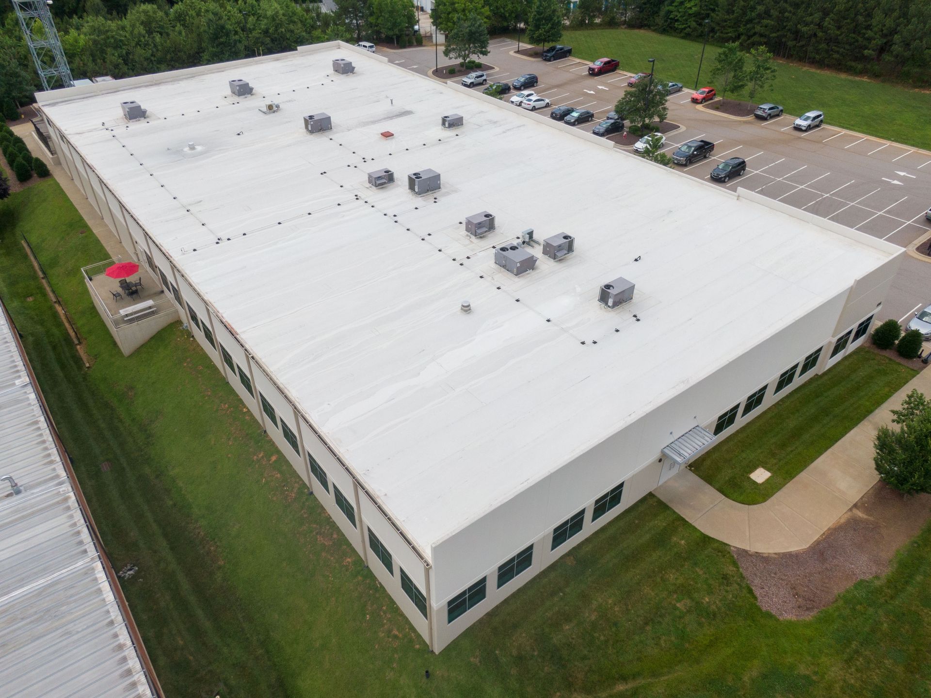 Aerial view of a large white industrial building with HVAC units on its roof, surrounded by green grass and a parking lot.