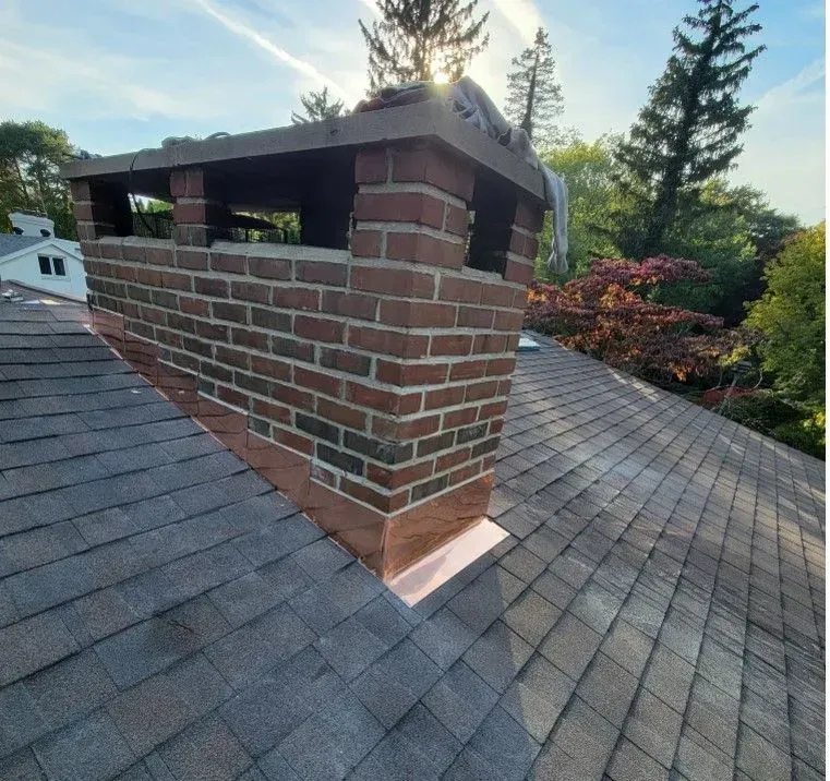 Brick chimney on a shingled roof with copper flashing, viewed from above, surrounded by trees.