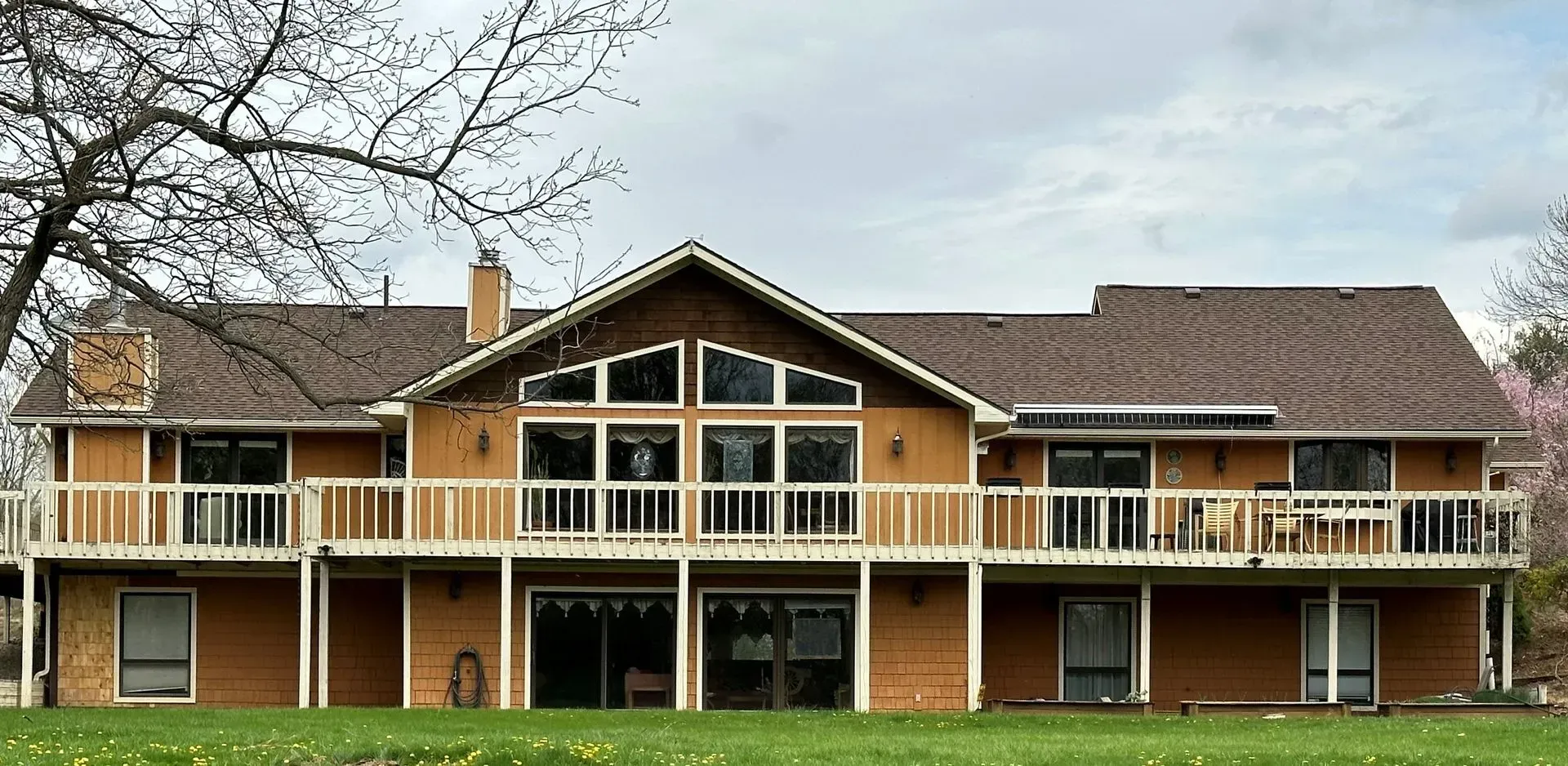 Large, tan house with a brown roof and white balconies. Green grass in the foreground; cloudy sky.