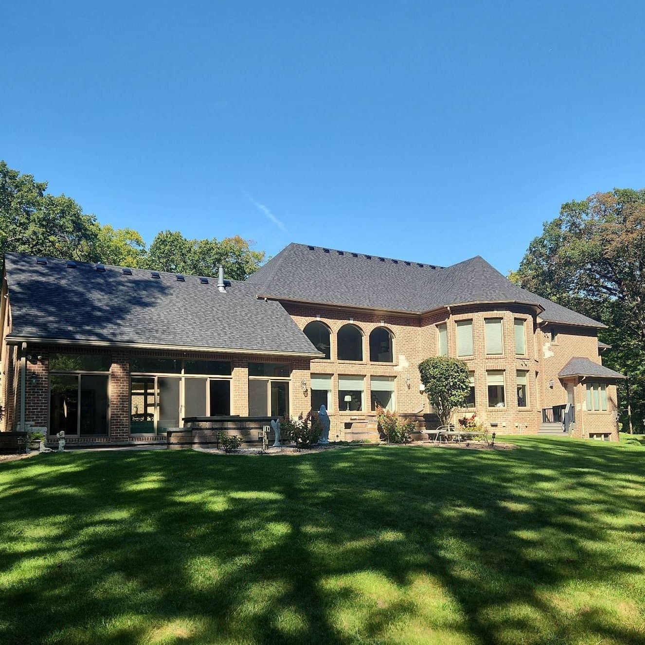 Large stone house with dark roof, surrounded by green lawn and trees, under a blue sky.