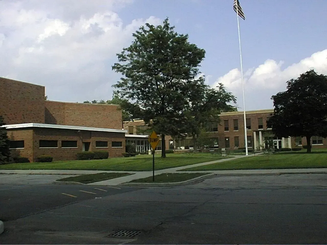 Brick school building with flag pole, trees, and green lawn on a sunny day.