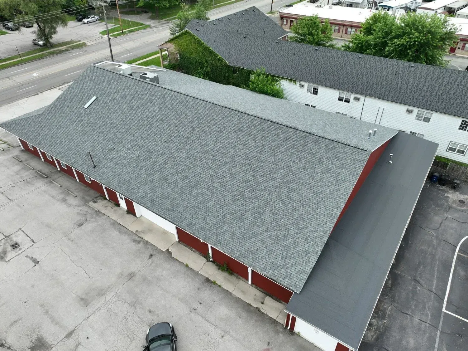 Aerial view of a long, red-sided building with a gray shingle roof, surrounded by pavement.