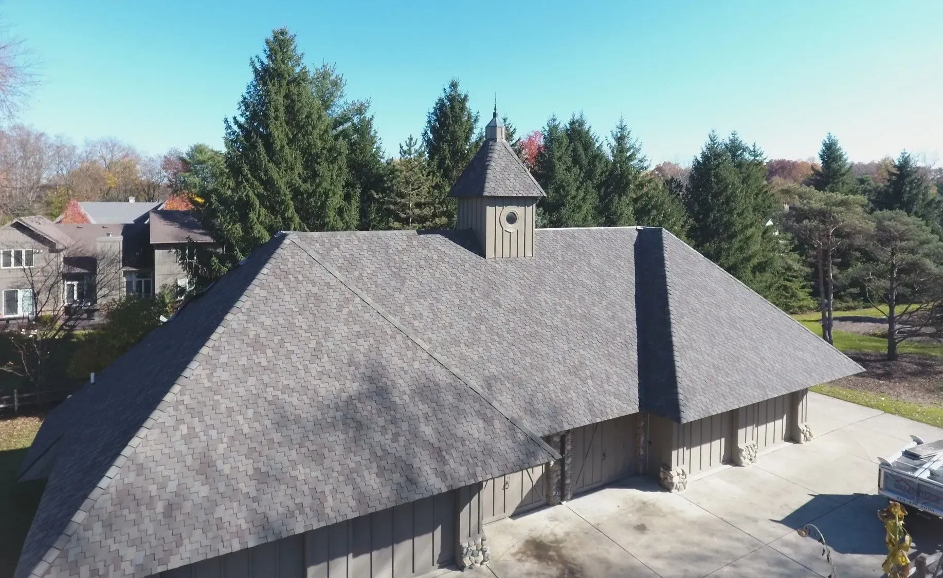 Gray shingled roof of a building with a tower, set against trees and a blue sky.
