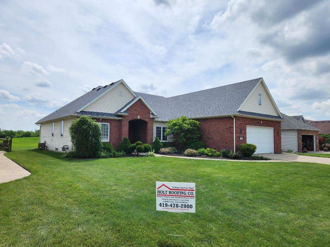 House with a new gray roof, brick facade, and green lawn on a sunny day. A sign is in the foreground.